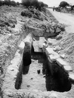 Excavated Tomb 13 at Amathus, a cut passage with large stone blocks, a dirt road and a parked car beyond.