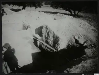 Excavation at Amathus with stones and trees; pale spoil heaps around a dark opening and a large block