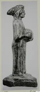 Limestone figurine of a standing woman holding a round object at her side, on a rectangular base, seen in profile.