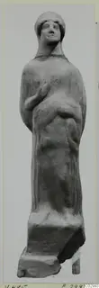 Terracotta figurine of a standing draped figure with a close-fitting head covering and one arm bent across the chest.