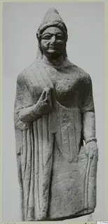 Standing limestone statue of a figure in a long robe with a conical headdress and one hand raised to the chest