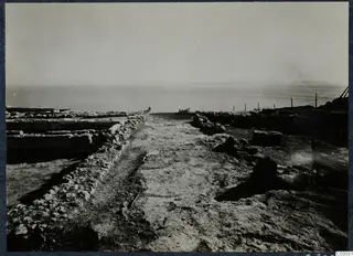 Dirt path between low ruined stone walls at Vouni palace, running toward the sea with a wire fence on the right.