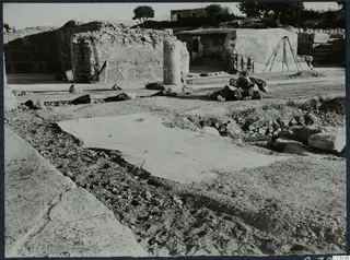 Ruined walls, a column drum and a heap of stones at the palace at Vouni, with trees and a house behind.