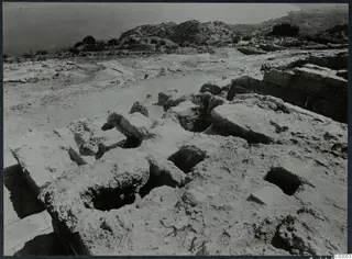Excavated stone walls and rectangular pits at Vouni, with a row of stakes along the edge and the sea and hills beyond.