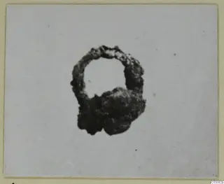 Heavily corroded iron object with an open loop at the top and a lumpy mass at its base, photographed on a card mount