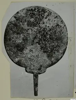 Corroded round metal disc with a narrow tang, photographed on a grey card mount, from Marion Tomb 41
