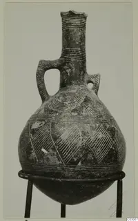 Pottery Red Polished III jug with a tall neck, loop handle and incised decoration, shown on a metal stand