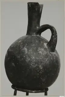 Dark round-bodied jug with a tall neck, a single loop handle and a pouring lip, standing on a metal stand.