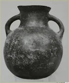 Pottery jug with two loop handles and a dark, mottled surface, standing on a pale base