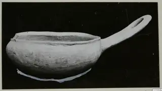 Plain white hand-made pottery bowl with a long handle, photographed against a dark background.