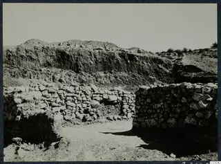 Stone wall foundations of a house at Mersinaki excavation, seen from the north, with a dug earth bank behind.