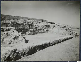 Stone wall foundations and a flat floor area at Vouni Palace, with two small figures standing on the ridge behind