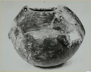 Globular pottery vessel with a wide mouth and two lugs at the shoulders, one pierced by a small hole, shown on a plain light background.