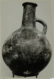 Pottery Red Polished III jug with a single loop handle, standing on a metal stand against a plain background