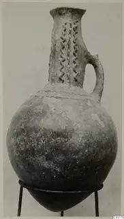 Red Polished III jug with a tall neck, applied wavy vertical strands and a single loop handle, shown on a metal stand.