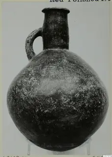 Red Polished III jug with a single loop handle and rounded body, shown on a small stand against a plain grey backdrop