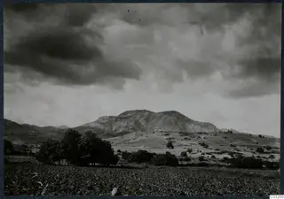 Dark, cloud-filled sky over a rocky hill with a dark tree cluster and a cultivated field in the foreground, at Soli.