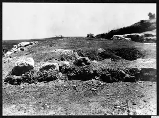Stony ground at Soli with large pale stones and earth mounds, a hillside with a tree beyond