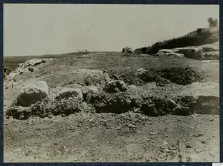 Theatre at Soli: stony ground with low earth mounds and rubble in the foreground, a scrubby hillside with a tree at right.