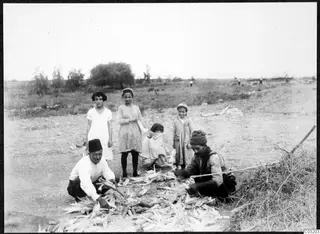 Two men shucking maize cobs on bare ground at Soli, with four girls and children standing and crouching nearby
