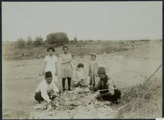 Two men shucking maize cobs on dry ground at Soli, with four children standing and crouching behind them.