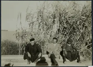 Three men seated on a wooden plank platform in front of tall reeds, Karavostassi, Soli.