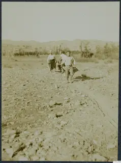 Three men walking across a stony field at Soli, with low hills and trees in the background.