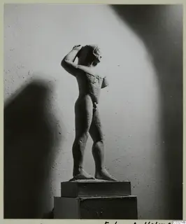 Limestone statue of a standing nude male figure with one arm raised to the head, photographed on a wooden pedestal