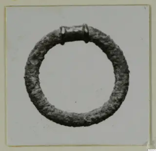 Corroded metal ring with a collar-like band and small loops at the top, photographed on a pale background.