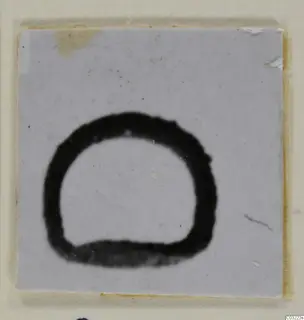 Small square of pale card or ceramic with a thick black ring-shaped mark, open at the bottom, on a cream backing