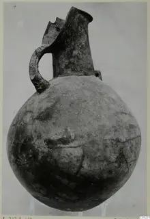 Red Polished III jug from the Vrysi tou Barba tombs at Lapithos, shown on a stand against a plain backdrop