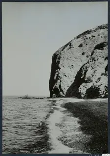 Steep pale rock cliff rising above a pebbly shore, with waves running in from the sea on the left.