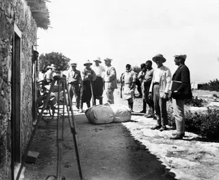Group of men standing in a row beside a stone building at Vouni, with a large rolled bundle on the ground