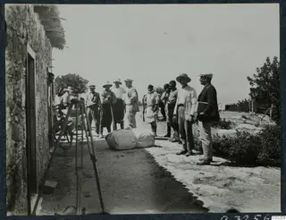 Men standing on a dirt track beside a stone building, with a rolled bundle on the ground and a bicycle against the wall at Vouni.