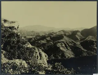 Landscape at Vouni: a view over hills and a mountain range from a rocky outcrop with bushes in the foreground.
