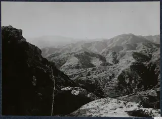 Mountain landscape seen from a rocky height at Vouni, with hazy sky, a dark cliff edge and scrub-covered slopes.