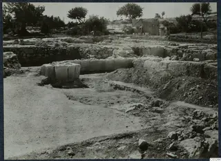 Excavated ruins of stone walls and blocks at the palace at Vouni, with trees and a tarpaulin shelter behind.