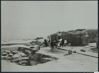 Workers repairing stonework in Room 47 at Vouni, with a standing man watching and the sea beyond the ruins.