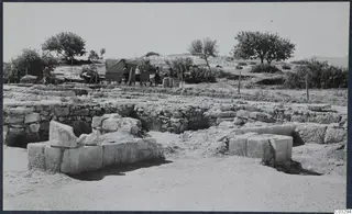 Excavated stone walls at the Vouni palace, with a canvas tent, three people and a wire fence behind