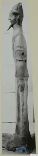 Tall terracotta standing figure in profile, with a conical headdress, long pointed nose and pointed beard, shown against a pale wall.