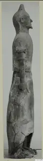 Tall terracotta statue of a standing figure in side profile, with a pointed head and cracked surface