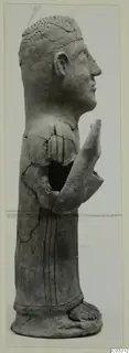 Side view of a grey terracotta standing figure with raised hands, cracked and partly broken.