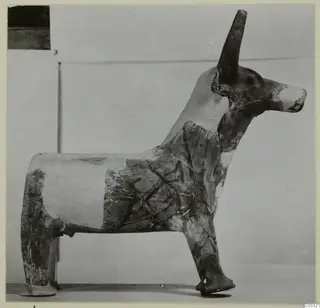 Terracotta figurine of a horned animal standing on four legs, with dark painted bands on a pale surface