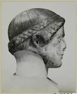 Carved stone head in profile, with a band of incised hair over the forehead and ear, on a plain pedestal.
