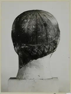 Back view of a carved stone head with grooved hair and a braided band, set on a pale neck support and plinth