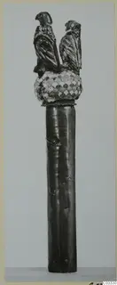 Upright ornamented object: a dark cylindrical shaft topped by a scale-patterned knob and two bird figures