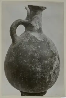 Pottery Red Polished III jug with a rounded body, tall neck, lipped spout and strap handle, standing on a dark base
