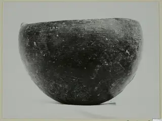 Rounded bowl with a wide mouth and dark, smooth surface with fine horizontal striations, photographed alone on a plain background