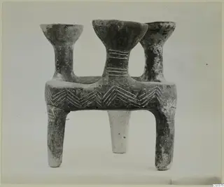 Grey pottery vessel with three flared mouths on a rectangular body and legs, with incised chevron decoration