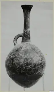 Pottery Red Polished II jug with a tall neck, flared rim and single loop handle, shown on a stand